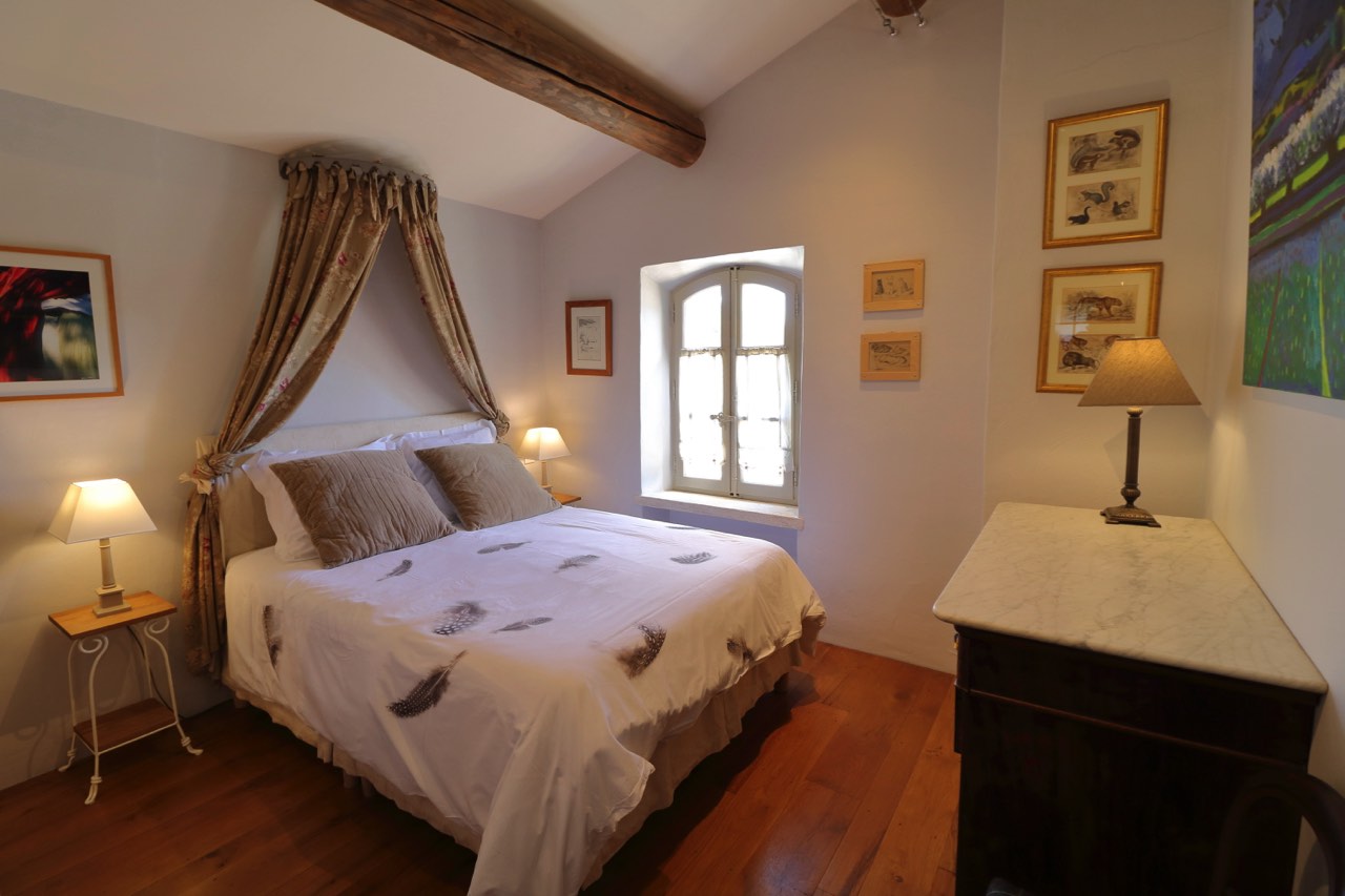 A cozy, well-lit bedroom featuring a large bed with a white feather-patterned duvet, wooden beams on the ceiling, framed artworks on the walls, and a window letting in natural light.